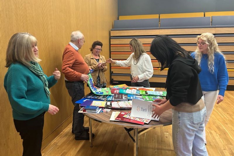 five women and one man gathered around a circular table covered in information booklets, with a wooden wall behind